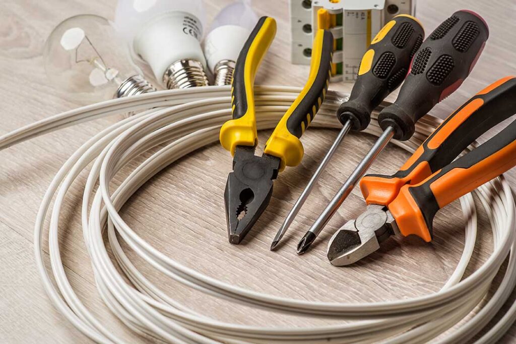 A hand plugging multiple electronics into a single wall outlet with visible power strips and extension cords.